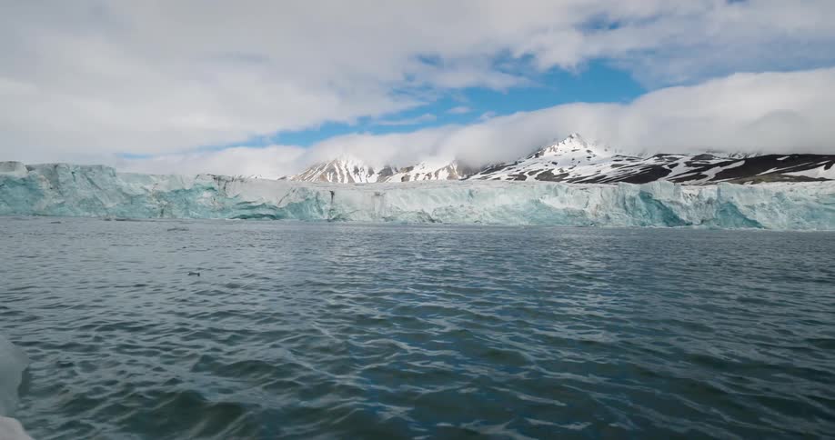 This footage showcases a vast blue glacier terminating in the ocean with snowy mountain peaks visible behind it under a cloudy sky the camera pans across the water where small chunks of ice float in the foreground near the massive ice wall