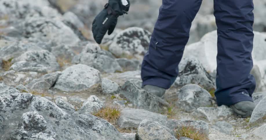 A close view shows a person wearing hiking boots and dark pants carefully walking across a steep rocky terrain