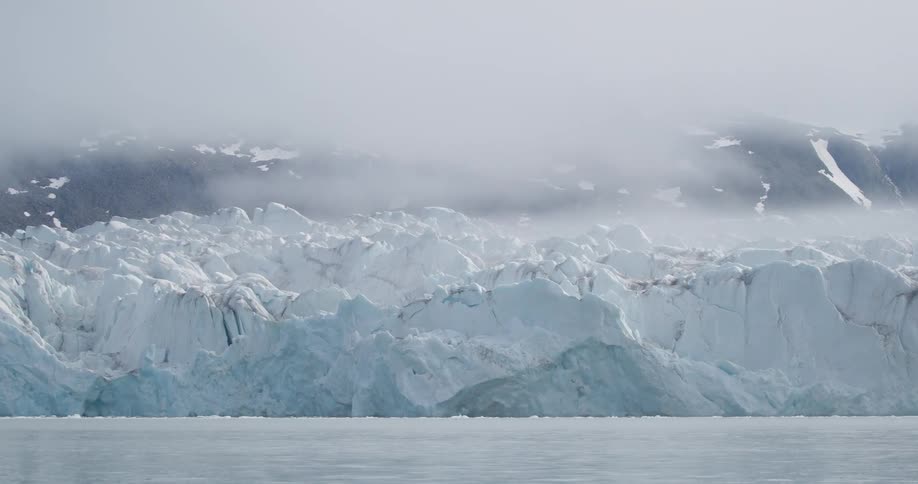 Glacier front with bluish ice blocks beside calm water and snowcovered mountains under fog in the background