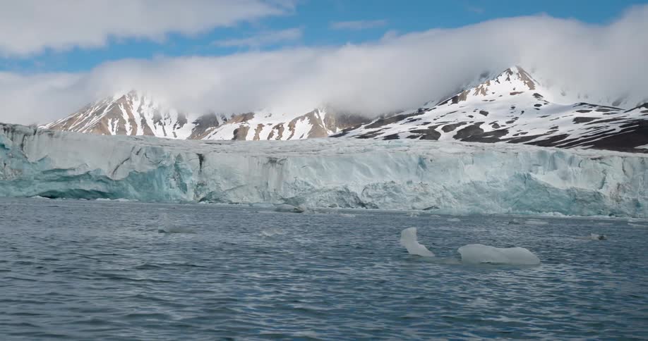 A wide shot shows a massive blue tinged glacier meeting the dark ocean water with snow covered mountains and low hanging clouds in the background
