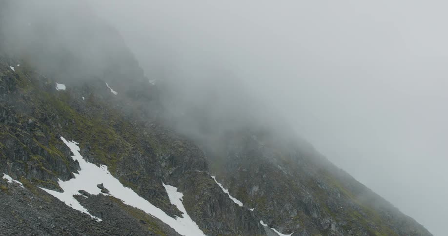 Thick gray clouds drift slowly across the steep rocky face of a mountainpatches of white snow cling to the dark mossy slopes beneath the heavy mist