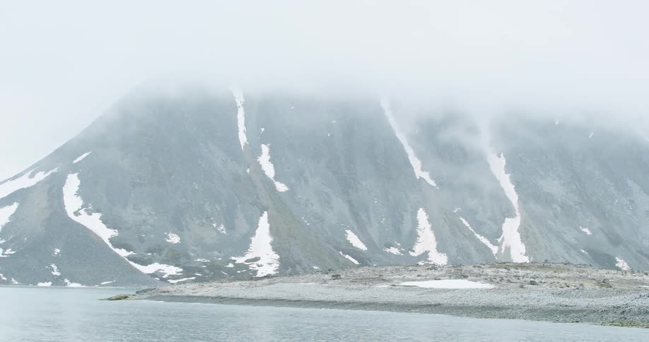 Steep mountain with patches of snow partially covered by fog beside a coastal shoreline