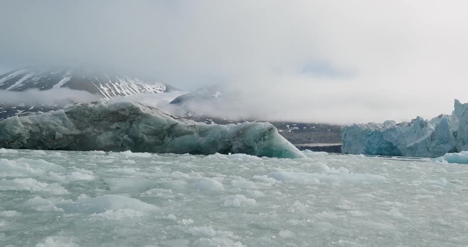 Fragmented ice covering the water surface with larger blocks near mountains partially shrouded by fog