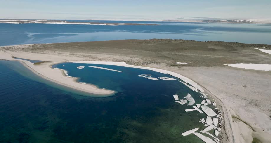 An aerial drone shot captures a vast arctic coastline where chunks of white ice float in deep blue water near a curved sandy spit and rocky tundra under a clear sky