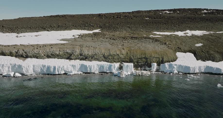 An aerial shot tracks slowly to the right along a rocky snowdusted shoreline where a shelf of white ice meets the clear dark water the rugged brown landscape above contrasts with the bright ice and the tealtinted depths of the calm sea