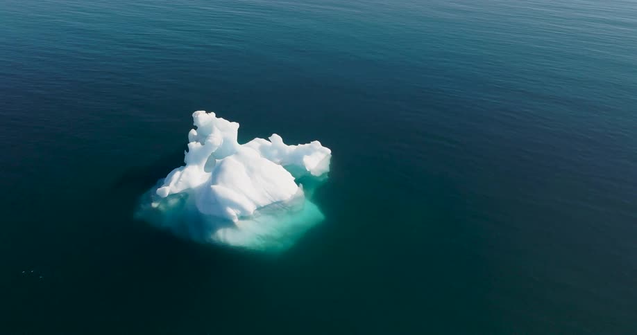 This video features a highangle drone shot of a jagged white iceberg drifting in deep blue water sunlight reflects off the surface of the sea highlighting the icebergs tealcolored submerged base
