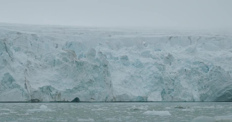 A massive ancientlooking glacier wall of jagged blue and white ice meets the grey arctic waterssmall chunks of ice float in the foreground as a thick overcast mist hangs over the frozen landscape