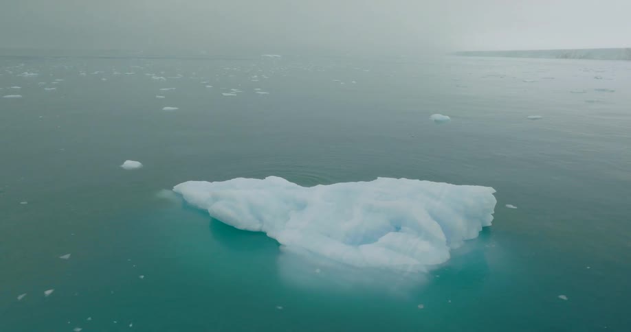 A large white iceberg floats in the center of vast turquoise arctic watersthe camera pans out to reveal a misty horizon filled with smaller ice fragments and a distant glacial shelf