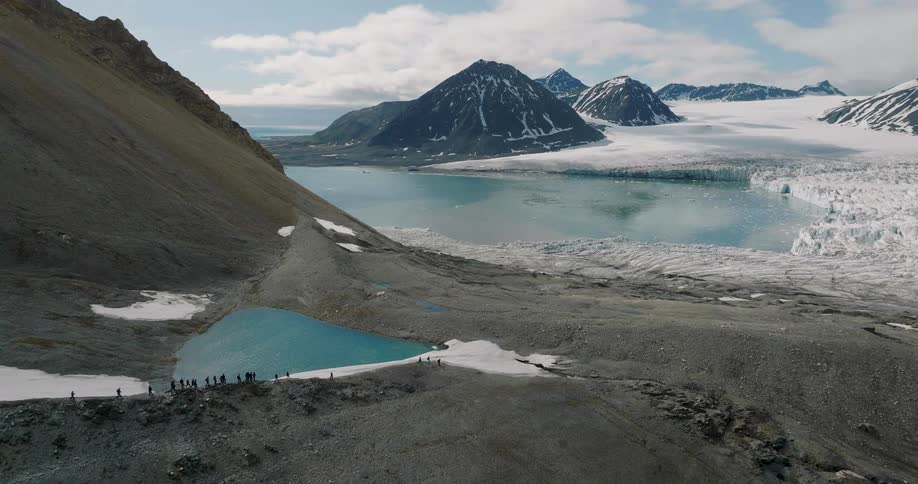 Hikers walk along a rocky ridge beside a turquoise glacial lake with mountains and ice fields in the distance