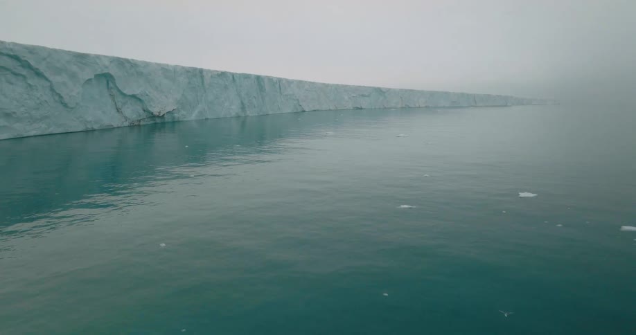 Continuous ice wall along calm ocean waters under overcast sky and low visibility