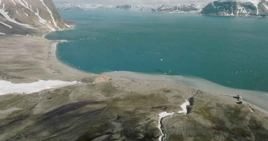 Aerial view of a wide arctic bay framed by rugged snow dusted mountains beside a sweeping shoreline
