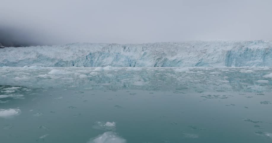 A massive wall of bluetinted glacial ice stretches across the horizon under a heavy overcast sky the surrounding water is filled with a dense field of floating ice chunks and slush that ripple as the camera moves forward