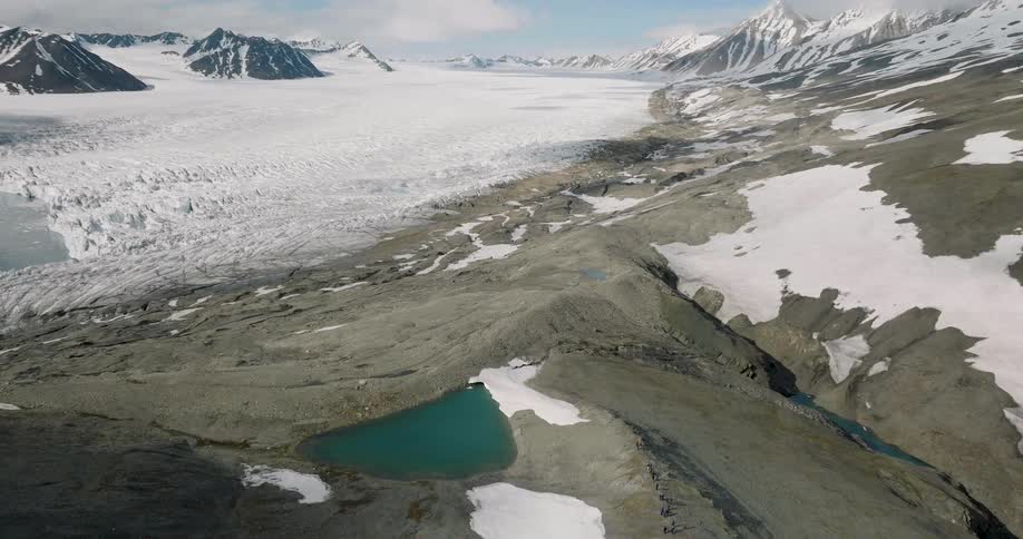 An aerial view shows a vast glacier meeting a turquoise lake in a rugged arctic landscape with snowy mountain peaks