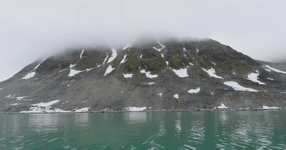 A lowangle shot from a boat moving past a large rocky mountain partially covered in snowthick mist and lowhanging clouds shroud the upper peaks of the mountain while the turquoise water ripples in the foreground