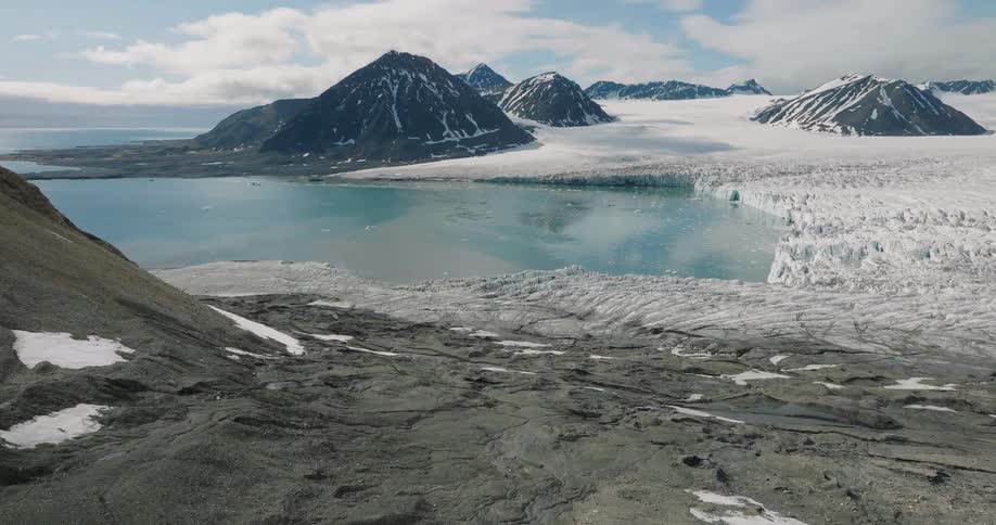 Glacier advancing toward a bluewater inlet surrounded by rocky mountains partially covered with snow