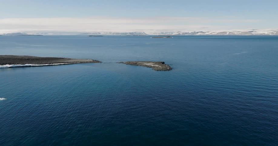 An aerial shot reveals a dark rocky peninsula extending into deep blue arctic watersin the far distance snowcapped mountains and icecovered plains sit beneath a clear bright sky