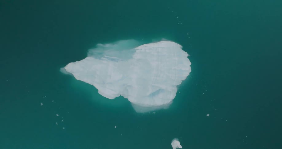 A top down aerial shot captures a single bright white iceberg drifting slowly in the middle of vast dark teal water