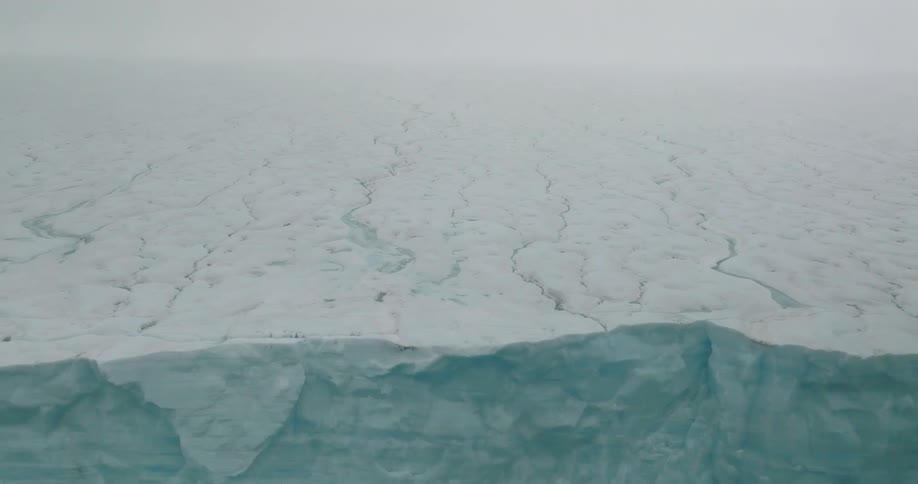 This aerial footage captures an expansive cracked ice sheet under a heavy overcast skythe camera pulls back to reveal the massive vertical face of a glacier where the ice meets dark open water