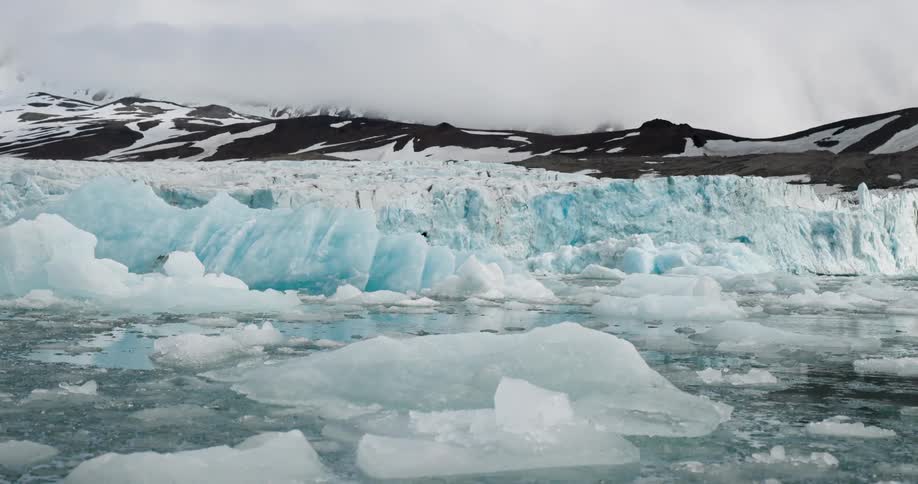 Glacier front with fragmented blue ice beside cold water and mountains partially covered by snow in the background