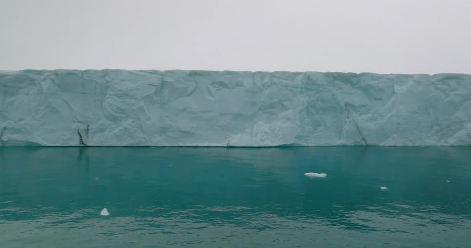 A towering icy glacier rises above calm arctic waters beneath a soft sky