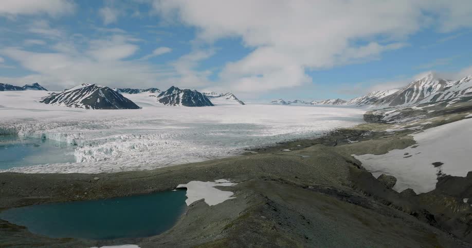 A wide aerial shot shows a massive white glacier flowing through a valley of dark rocky mountains under a blue sky with scattered clouds