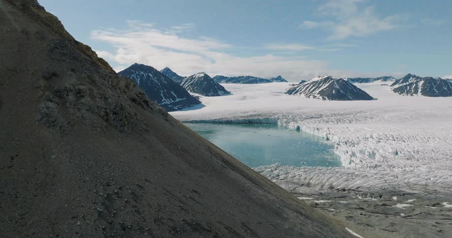 A steep rocky mountainside reveals a vast glacier and turquoise lake surrounded by snow capped peaks