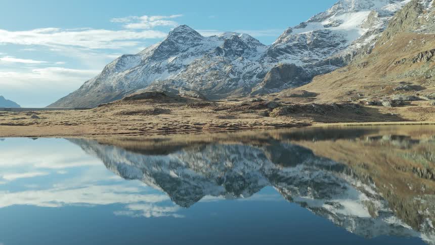 Snow capped mountains reflect on the surface of a still alpine lake under a clear blue sky while dry grass and rocky terrain border the water edge in a vast natural landscape