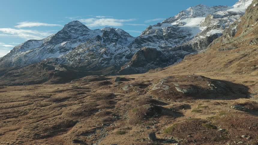 A drone flies low over a wide rocky valley filled with brown grass and small boulders moving toward a massive range of snowcapped mountains under a bright clear blue sky during a sunny day