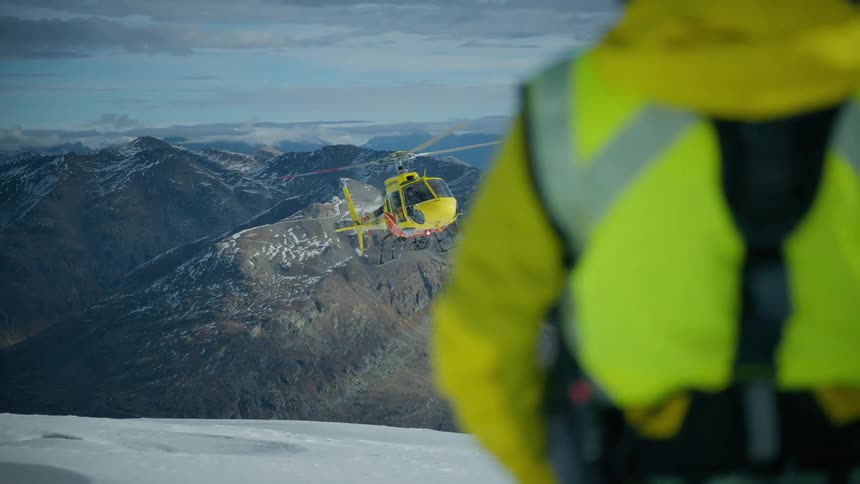 Man stands on a snow covered mountain watching an approaching helicopter
