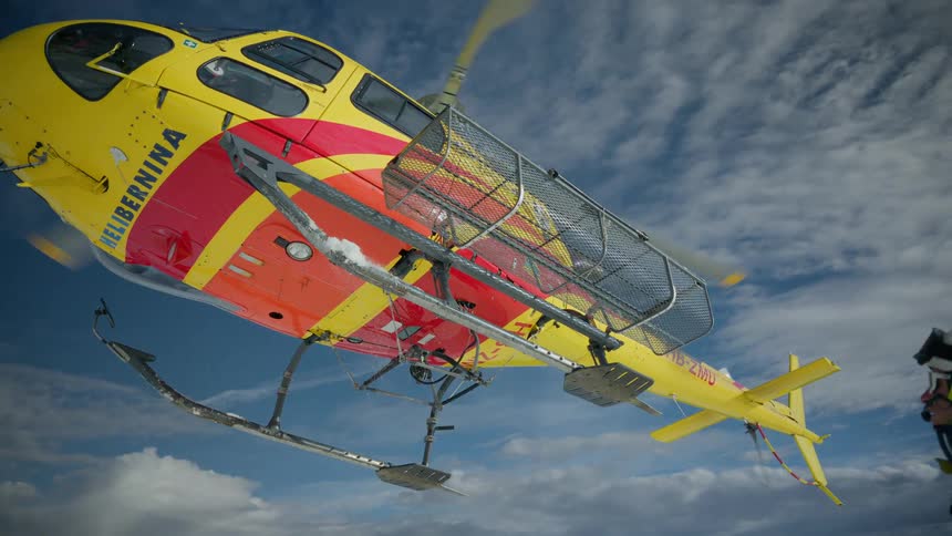 A yellow and red rescue helicopter takes off from a snowy mountain peak while a person in safety gear watches from the ground