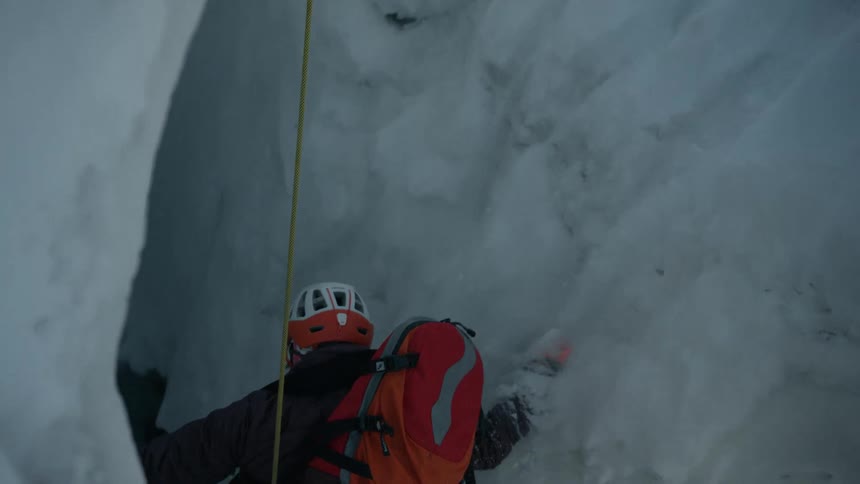 A climber wearing an orange backpack and helmet falling into a crevasse while attached to a yellow safety rope during a cold mountaineering expedition