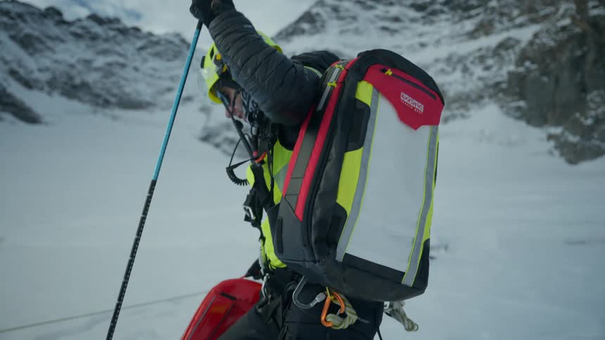 A person wearing a mountain rescue uniform and a large technical backpack stands on a snowy slope and uses a long probe to check the snow depth