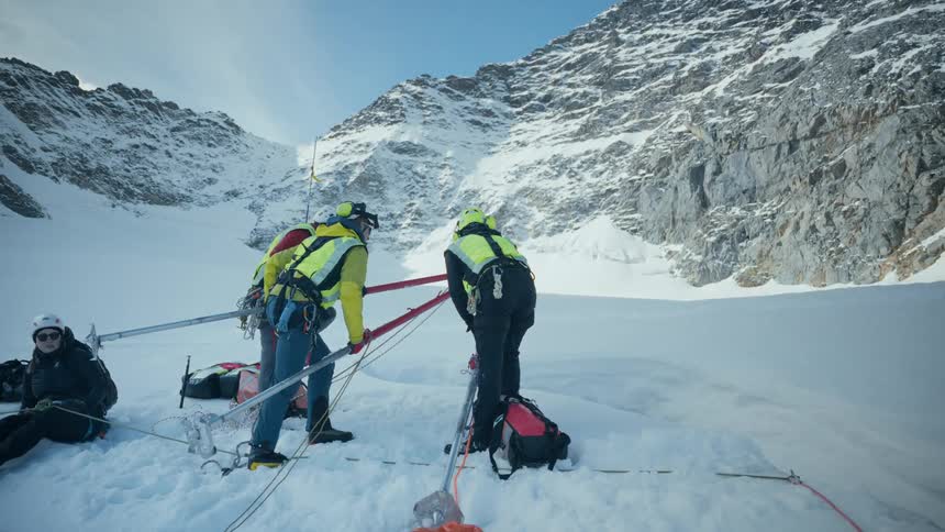 A mountain rescue team in highvisibility gear works with a specialized tripod and pulley system to perform a recovery or rescue on a snowy glacierthe team operates at the base of a steep rugged mountain face carefully managing ropes and equipment in
