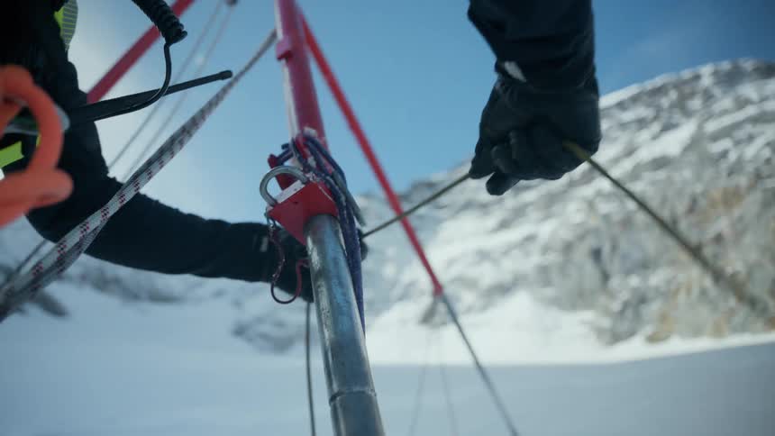 A mountain rescue or research team operates a red tripod hoist system on a snowy highaltitude slope the footage captures a closeup of a person in technical gear carefully managing ropes and clips against a backdrop of jagged sunlit peaks