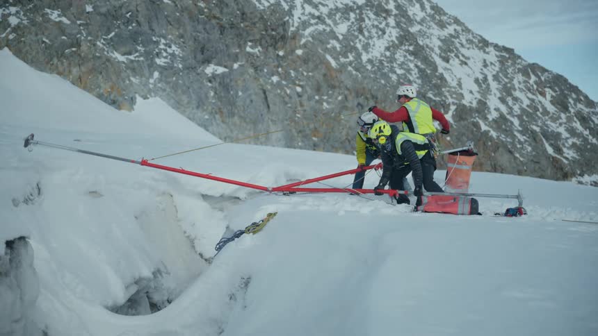 A mountain rescue team works in unison to deploy a mechanical winch carefully extending its long red arm over the gaping maw of a deep crevasse in a snowy landscape