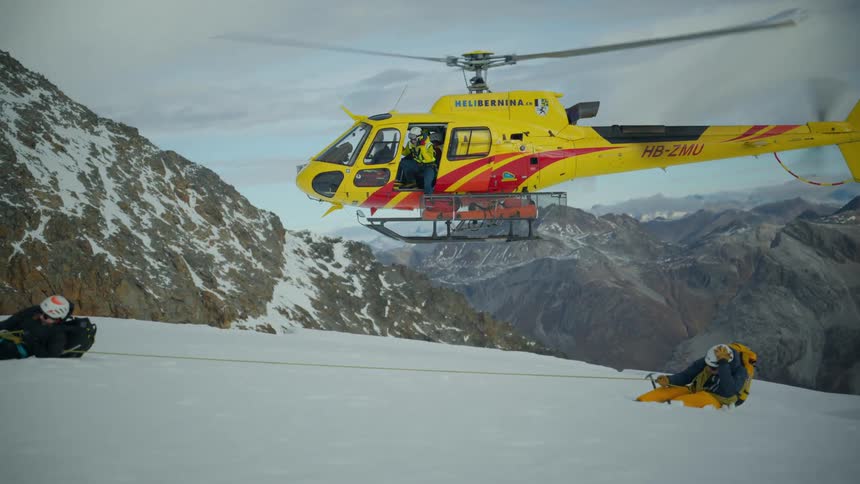 A yellow rescue helicopter lands on a snowy mountain ridge next to two tethered climbers