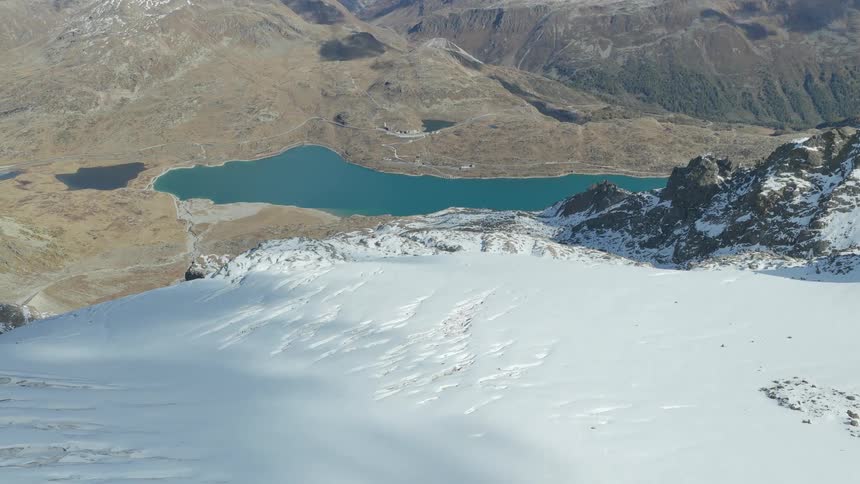 From an aerial perspective the camera glides over a pristine snowcovered mountain peak to reveal a vibrant turquoise lake nestled deep within a rugged sunlit valley below