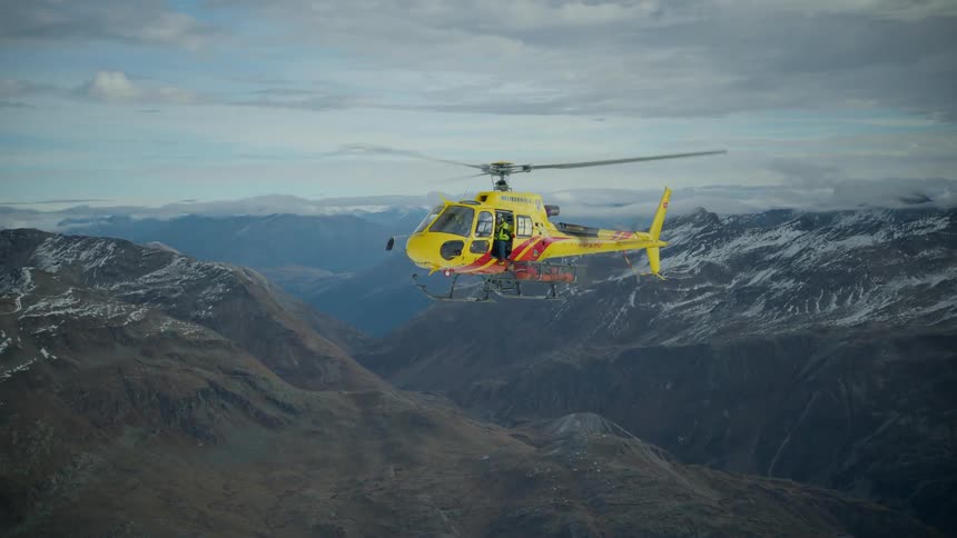A yellow rescue helicopter flies steadily through a vast mountain range under a cloudy skythe aircraft moves across a deep valley with snowdusted peaks visible in the background