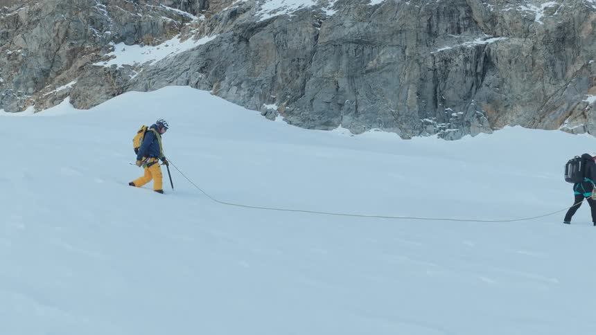 Two mountain climbers wearing technical gear and backpacks walk across a steep snow covered slope while connected by a long safety rope