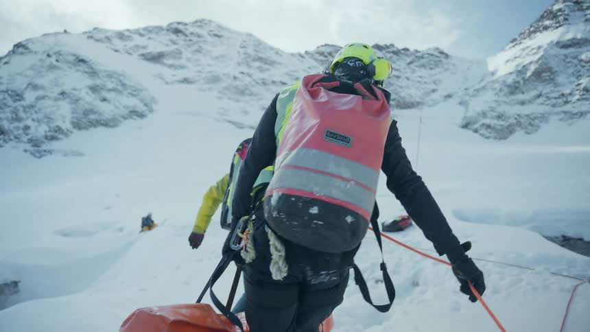 People follow a guide rope across the snow while carrying bags toward a woman sitting and holding a fishing pole on a winter day