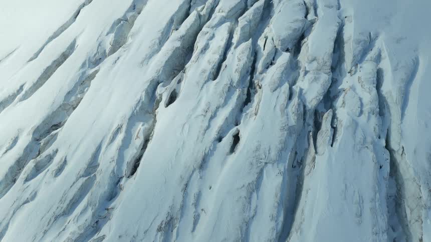 A topdown aerial view drifts across the deeply fissured surface of a vast mountain glacier revealing jagged crevasses and the stark texture of the ice