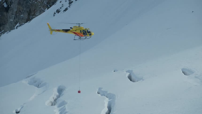 A yellow helicopter flies across a snowy mountain landscape while carrying a red cargo basket suspended by a long cablethe aircraft maneuvers over a series of deep icy crevasses as it transports the load through the alpine environment