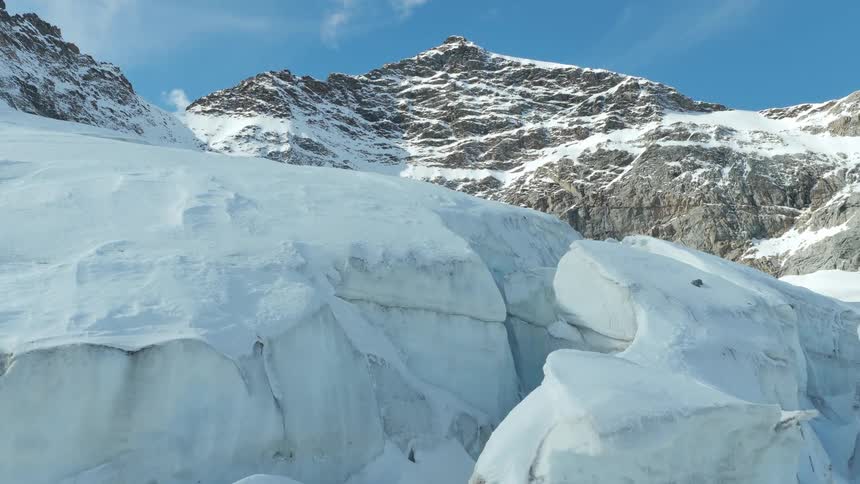 A camera moves slowly over a massive fractured glacier revealing deep blue cracks and jagged ice blocks in the foreground while a steep rocky mountain peak rises against a clear sky in the background