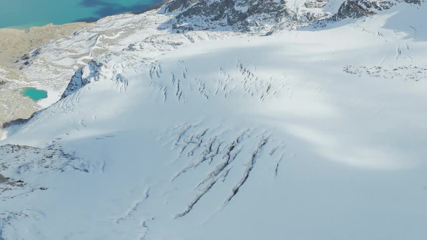 An aerial drone shot pans slowly over a vast snowcovered mountain range revealing deep cracks in a glacier while a bright blue alpine lake sits nestled among rocky cliffs in the distant background