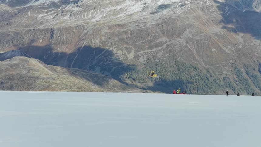 A yellow and red rescue helicopter flies over a vast white snowy plain toward a group of people and equipment on the ground with a large rocky mountain in the background