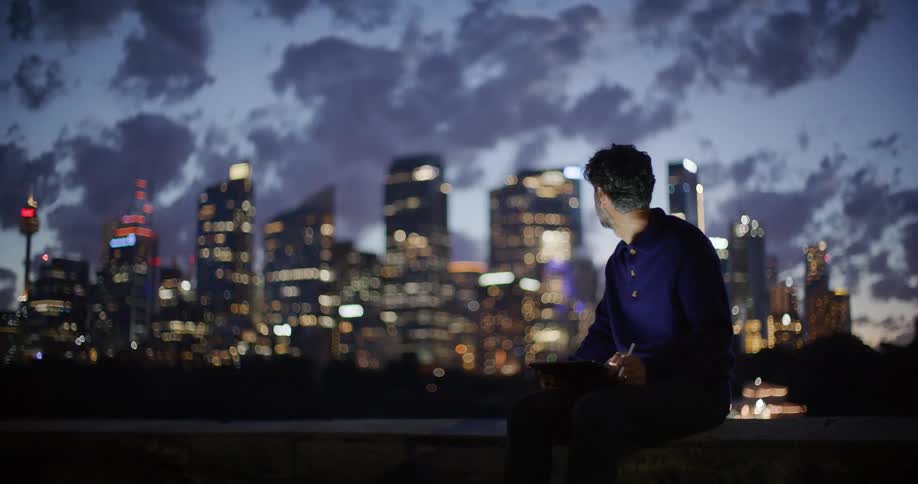 Man sitting on the roof of a building with a tablet computer as he looks at towers and skyscrapers in the distance