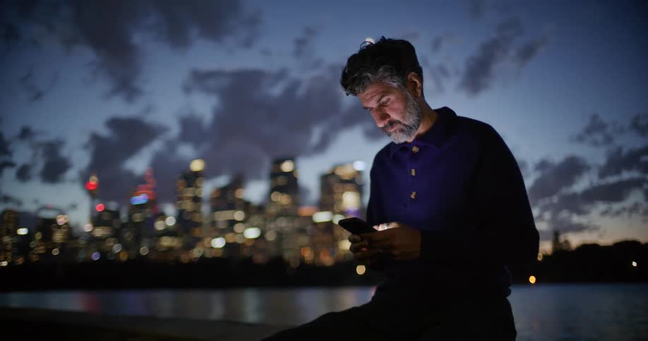 A middleaged man with a grey beard sits on a stone ledge at night looking down at his glowing smartphone screen while a blurred city skyline and harbor lights illuminate the dark background behind him