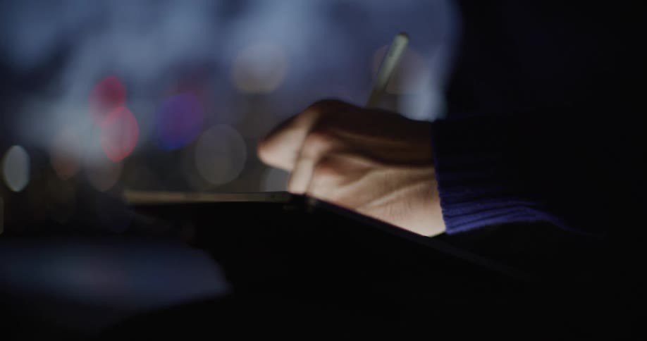 Hand uses a digital stylus on a tablet in a lowlight environment with blurred city lights