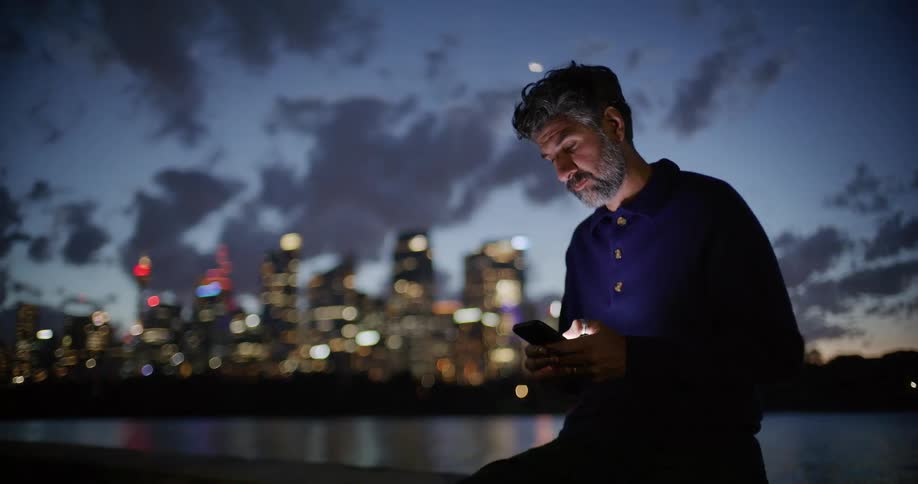 A middleaged man with graying hair sits on a waterfront ledge at night looking at a brightly lit city skyline before turning his attention back to his glowing smartphone screen while clouds drift across the dark sky behind him