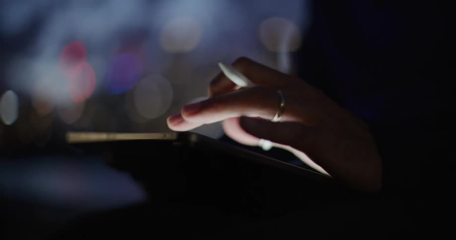 A closeup view shows a persons hand wearing a ring and holding a white stylus while interacting with a tablet screenthe scene is set at night with a blurred background of city lights creating a soft bokeh effect